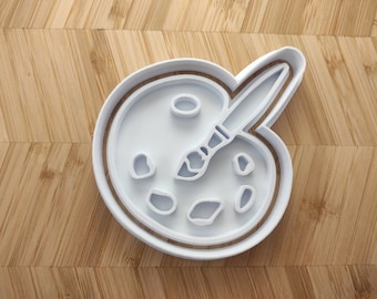 May include: White plastic cookie cutter shaped like a paint palette with a paintbrush. The palette has five paint wells.