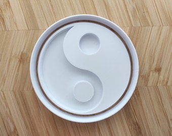 Może przedstawiać: Biała plastikowa foremka do ciasteczek w kształcie symbolu yin yang. Foremka ma podniesiony brzeg wokół zewnętrznej krawędzi.