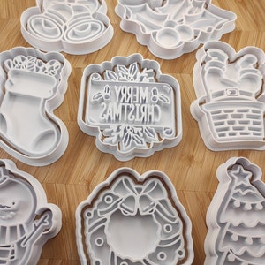 May include: A set of ten white plastic Christmas cookie cutters. The cutters are shaped like a stocking, a snowman, a wreath, a Christmas tree, a Santa Claus, a bell, a candy cane, a gingerbread man, a star, and a reindeer.