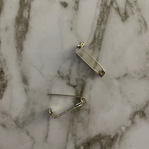 May include: Two silver-toned safety pin closures for jewelry making. The pins are closed and lying on a white marble surface.