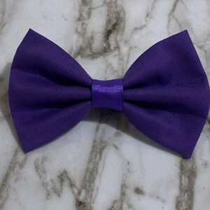 May include: A solid purple bow tie made of fabric. The bow tie has a center band of a slightly darker purple color. The bow tie is a classic accessory for formal wear or adding a touch of style to any outfit.