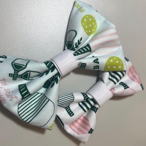 May include: Two white fabric bow ties with a repeating pattern of pickleball paddles, balls, and text in green, pink, and yellow. The bows have a white band in the center.