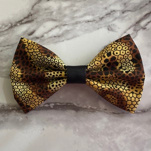 May include: A brown and gold bow tie with a geometric pattern. The bow tie is made of fabric and has a black adjustable strap.