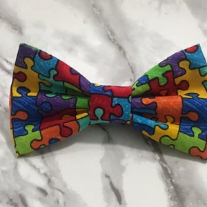 May include: A colorful bow tie featuring a pattern of interlocking puzzle pieces in various colors, including red, blue, green, yellow, and purple. The bow tie is a vibrant accessory.