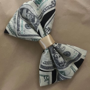 May include: A fabric bow tie featuring a repeating print of United States five-dollar bills in shades of green, black, and white. The center of the bow is cinched with a smooth, solid light beige ribbon band, creating a gathered effect.
