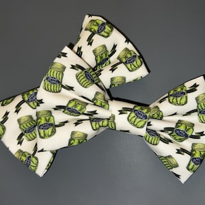 Op de afbeelding: Een witte vlinderdas met een herhaald patroon van groene augurkenpotten. De potten hebben blauwe en witte etiketten en zijn geplaatst op een zwart lintontwerp. Een klassiek accessoire.