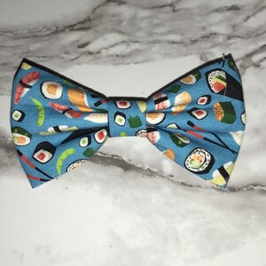 Può includere: Un papillon blu con un motivo di sushi. Il papillon presenta diversi tipi di sushi, tra cui maki, nigiri e sashimi.