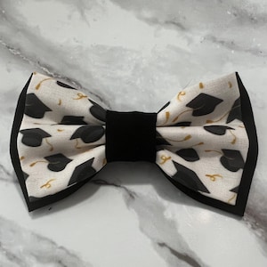 May include: A black bow tie with a white fabric pattern of graduation caps and gold tassels.