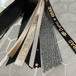 May include: A set of graduation tassel streamers in black, white, gold, and silver with gold lettering that says "GRAD" and "Congrats".
