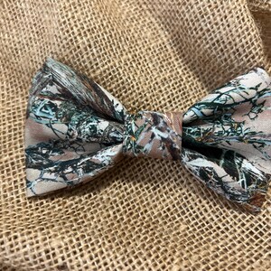 Può includere: Un papillon di camuffamento marrone e verde. Il papillon è fatto di tessuto e ha un motivo boschivo.