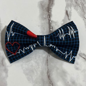 May include: A bow tie featuring a dark blue grid pattern overlaid with white EKG heart rhythm lines and small red heart icons. The fabric has a structured, folded design with a central knot, displaying a medical-themed print across the surface.