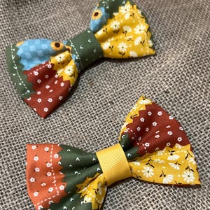 May include: Two handmade bow ties with a patchwork design. One bow tie features a blue, sunflower, and brown pattern, while the other has an orange, brown, and yellow floral pattern. Both have a green centre and are set against a burlap background.