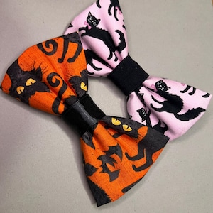 May include: Two fabric bow ties with a black cat pattern. One bow tie is orange with black cats and the other is pink with black cats.