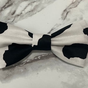 May include: A black and white cow print bow tie. The bow tie is made of fabric and has a classic bow shape.
