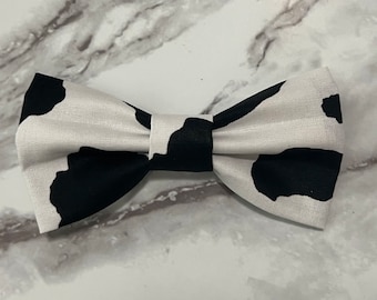 May include: A black and white cow print bow tie. The bow tie is made of fabric and has a classic bow shape.