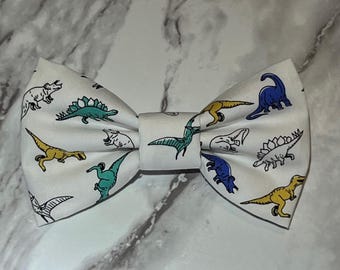 May include: White bow tie with a dinosaur pattern. The bow tie features various dinosaurs in blue, green, and yellow on a white background.