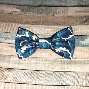 May include: A blue bow tie featuring a white and light blue wave pattern. The bow tie is set against a wooden backdrop with shades of blue, grey, and brown. A classic accessory for formal or semi-formal attire.