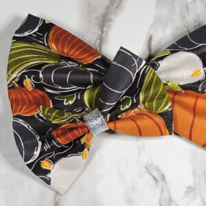 Puede incluir: Un lazo de tela con un estampado de calabazas otoñales. El lazo presenta calabazas naranjas, blancas y verdes sobre un fondo negro. Una banda plateada adorna el centro del lazo. Este accesorio es adecuado para diversas ocasiones.