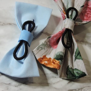 May include: Two fabric bow ties, one light blue and one with a floral pattern. Both bow ties have black elastic bands.
