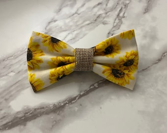 May include: A bow tie featuring a repeating sunflower pattern with yellow petals and dark brown centers on a cream-colored fabric. The center of the bow tie is cinched with a band of textured, light brown woven burlap material.