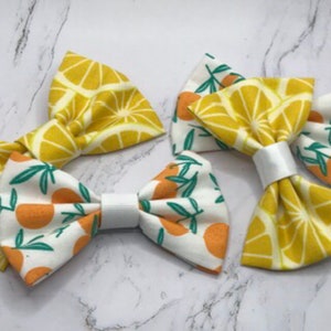 May include: Three fabric hair bows with a lemon and orange print. The bows are yellow, white, and green.