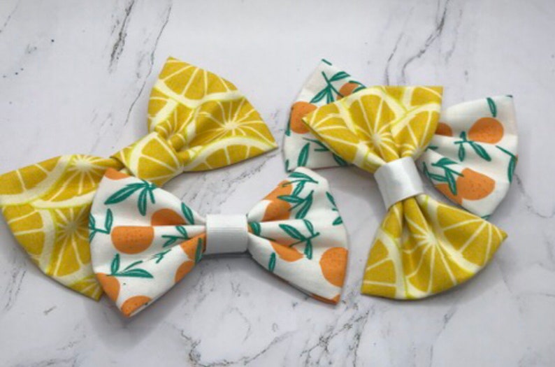 May include: Three fabric hair bows with a lemon and orange print. The bows are yellow, white, and green.