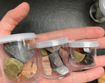 May include: Three small, clear plastic containers filled with various colorful rocks and minerals. The containers have white lids and are held in a person's hand. The rocks include shades of green, brown, gray, and red.
