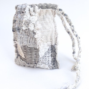 May include: A small, white drawstring bag made of patchwork fabric with a frayed edge. The bag is made of different shades of white and gray fabric and has a simple, rustic look.