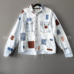 Peut inclure: Chemise en jean bleu clair avec des détails de patchwork. La chemise présente des patchs marron, bleus et à motifs cousus sur le devant, le dos et les manches. Elle a deux poches de poitrine et une fermeture boutonnée. La chemise est suspendue à un cintre noir.