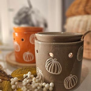 May include: Two ceramic wax warmers with pumpkin designs. The warmer on the left is orange with white pumpkins and the warmer on the right is brown with white pumpkins.