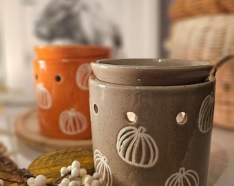 May include: Two ceramic wax warmers with pumpkin designs. The warmer on the left is orange with white pumpkins and the warmer on the right is brown with white pumpkins.