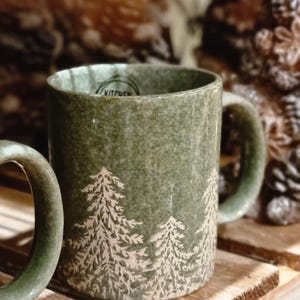May include: Green ceramic mug with white fir tree designs. The mug has a rounded handle and a speckled texture. The word "KITCHEN" is visible on the rim. The mug is sitting on a wooden surface.