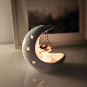 May include: White ceramic moon-shaped candle holder with a small metal bowl hanging from the crescent moon. The bowl is designed to hold wax melts or essential oils. A small white tea light candle is lit inside the holder.