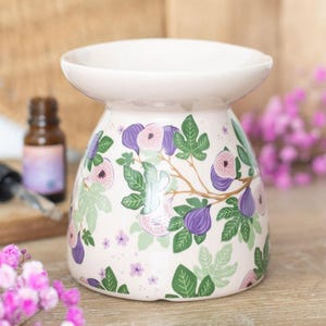 May include: A pale pink ceramic essential oil diffuser decorated with a fig pattern. The design includes purple figs, green leaves, and pink flowers. The diffuser has a wide, shallow bowl at the top for oil and water.