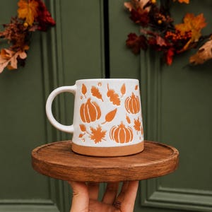 May include: A white ceramic mug with a handle, featuring an embossed pattern of orange pumpkins and autumn leaves. The base of the mug is unglazed terracotta, and the body has a smooth white finish with the orange design recessed into the surface.