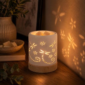 May include: A cylindrical white ceramic wax warmer featuring intricate cutouts of dragonflies and flowers. The warmer sits on a light-coloured wooden base and emits a warm glow, casting detailed dragonfly and floral shadows onto the wall behind it.