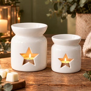 May include: Two white ceramic wax melt warmers with a glossy finish, each featuring a star-shaped cutout on the front. A lit tea light candle is visible inside each warmer, and a small white wax melt cube sits on a wooden board nearby.