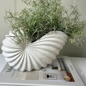 May include: A white, ceramic vase in the shape of a seashell, filled with green foliage. The vase is placed on a book titled "Minimalista". The vase has a spiral design and is a decorative home accent.