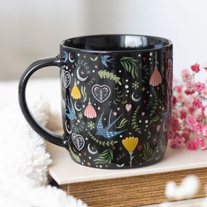 May include: Black ceramic mug decorated with a pattern of blue swallows, hearts, moons, and floral motifs in yellow, pink, and green. The mug has a black handle and interior, suitable for hot beverages.