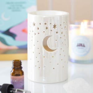 May include: A white ceramic essential oil diffuser with a crescent moon cutout and star-shaped perforations. The diffuser is cylindrical and has a glossy finish. Other items in the image include a bottle of essential oil and a lit candle.