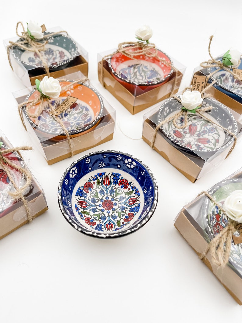 May include: Assortment of small, decorative ceramic bowls with intricate floral patterns in various colors, including blue, red, orange, and green. The bowls are packaged in clear plastic boxes with brown cardboard lids, tied with twine and decorated with small white flowers.