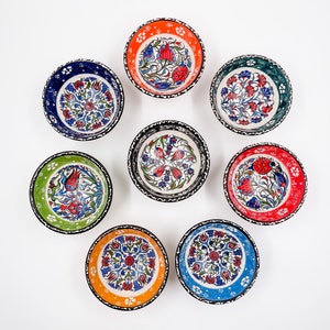 May include: Set of eight small ceramic bowls with colorful floral patterns. The bowls have a white background with blue, red, green, and orange accents. The bowls are arranged in a circle on a white surface.