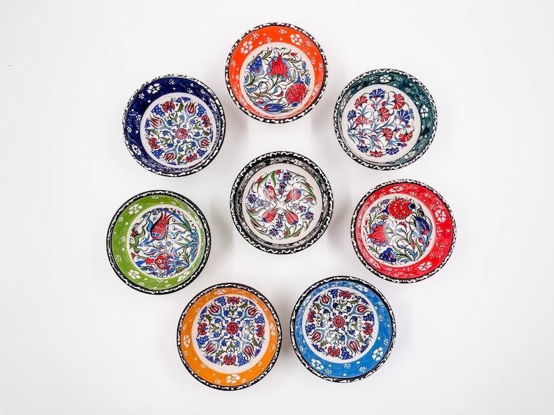 May include: Set of eight small ceramic bowls with colorful floral patterns. The bowls have a white background with blue, red, green, and orange accents. The bowls are arranged in a circle on a white surface.