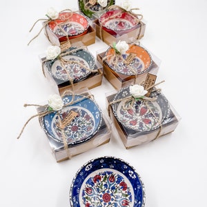 May include: Assortment of small, colorful ceramic bowls with floral patterns, packaged in clear boxes with brown lids and tied with twine. Each box includes a small tag with the text "Melissa & David 08.08.2021 Wedding Favors".