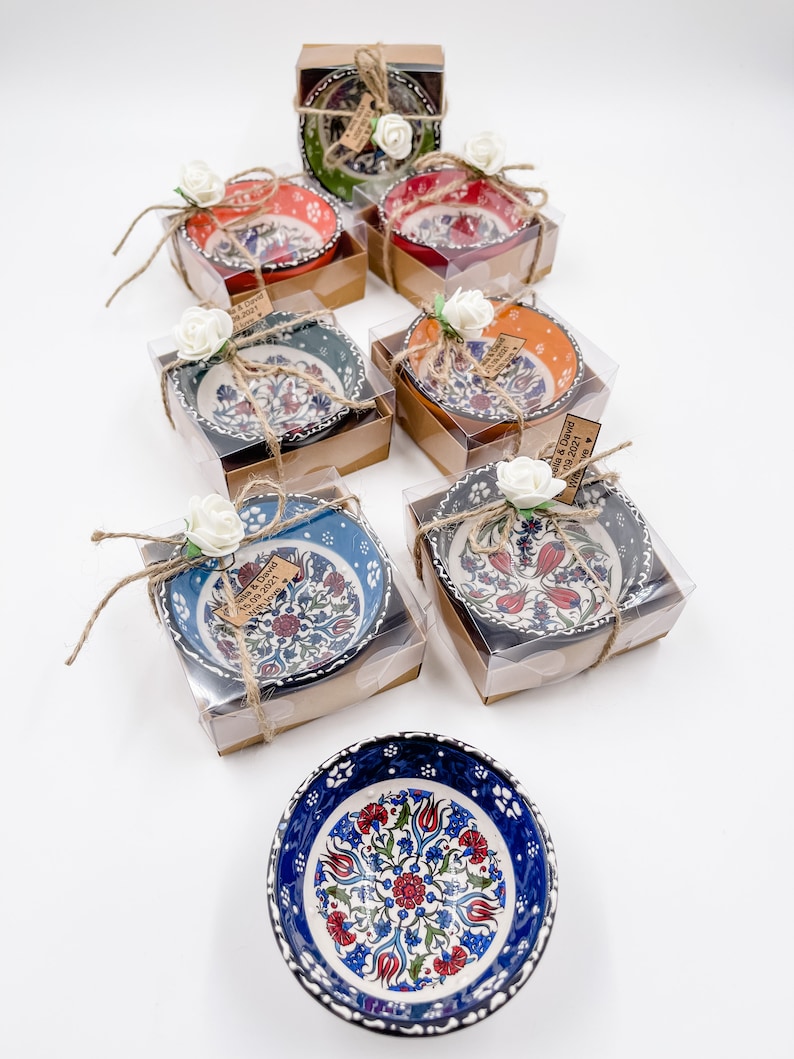 May include: Assortment of small, colorful ceramic bowls with floral patterns, packaged in clear boxes with brown lids and tied with twine. Each box includes a small tag with the text "Melissa & David 08.08.2021 Wedding Favors".