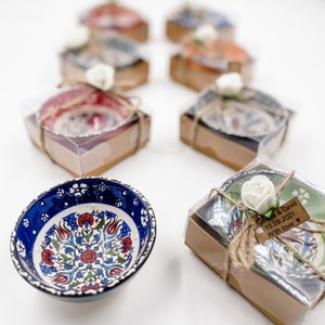 May include: A set of small, decorative bowls with intricate floral patterns in blue, red, and green. The bowls are packaged in individual boxes with a brown paper exterior and a clear plastic lid. Each box is tied with twine and a small white flower. One box has a wooden tag with the text "Sarah & David 15.09.2021 With love".