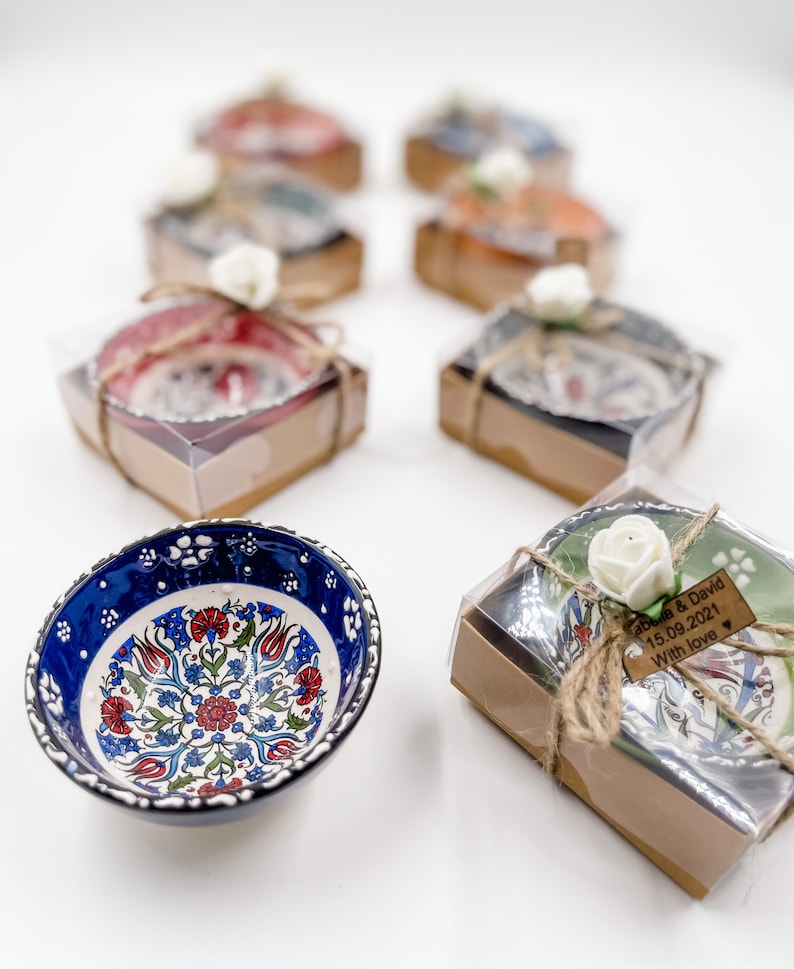 May include: A set of small, decorative bowls with intricate floral patterns in blue, red, and green. The bowls are packaged in individual boxes with a brown paper exterior and a clear plastic lid. Each box is tied with twine and a small white flower. One box has a wooden tag with the text "Sarah & David 15.09.2021 With love".