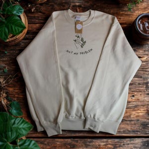 Può includere: Felpa color crema con la scritta "NOT MY PROBLEM" in nero. Il design include una mano che fa il segno della pace con foglie verdi. Un'etichetta è appesa al colletto.