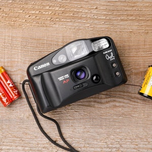 May include: A black Canon Sure Shot Owl Date 35mm AF camera with a lens, flash, and strap. Two red and gold AA batteries and a roll of Funtime 200 film are next to the camera. The items are on a wooden surface.