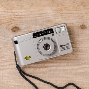 Op de afbeelding: Een zilverkleurige Pentax IQZoom 105 compactcamera voor film met een 35-105mm zoomlens, ingebouwde flitser en een zwarte polsband. De voorzijde bevat de tekst Pentax, IQZoom 105, Pentax Zoom 35-105mm Quartz Date en een gele Panorama-sticker.
